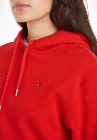 Hoodie en coton rouge avec une capuche à cordon de serrage, patch logo sur la poitrine et poignets côtelés. Le tissu a une texture douce et lisse.