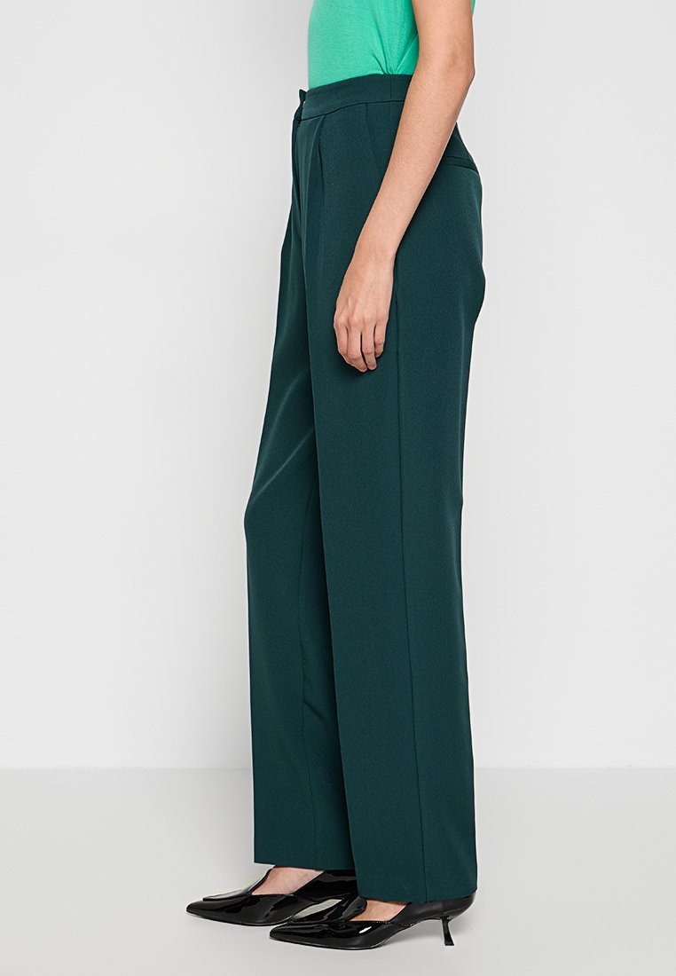 Lange, rechte broek in donkergroene stof, met een hoge taille en voorplooien. Gecombineerd met zwarte puntige schoenen.