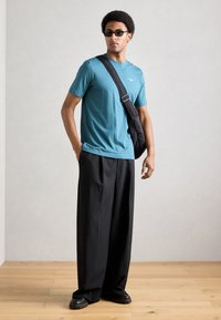 Tricou turcoaz, croială lejeră, asortat cu pantaloni negri cu picioare evazate. Modelul poartă ochelari de soare negri și are o geantă neagră tip crossbody.
