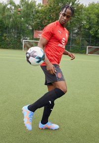 Maglia rossa con un motivo geometrico, pantaloni corti neri con logo e dettagli di design, calzini neri e scarpini blu con dettagli a zigzag arancioni.