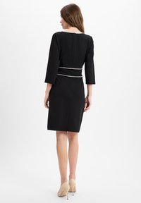 Schwarzes Kleid mit Dreiviertelärmeln, taillierte Form, betonte Taille mit einem weißen Band, Reißverschluss hinten, getragen mit nude-farbenen High Heels.