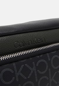 Musta teksturoitu nahkapinta, jossa on kohokuvioitu "Calvin Klein" -logo hopea vetoketjun yläpuolella kirjailtulla kankaan pinnalla.