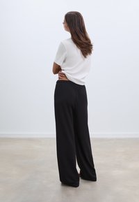 Witte top met korte mouwen en zwarte wijde broek. Gladde stof, ontspannen pasvorm. De broek heeft een elastische tailleband. Model staat met de rug naar de kijker.
