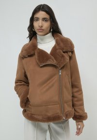 Giacca in finta pelliccia shearling marrone con ampio colletto, cerniera frontale e due tasche laterali; texture morbida, con una rifinitura in pelliccia morbida sui polsini e sull'orlo.
