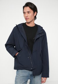 Fiatal férfi sötétkék kapucnis dzsekiben, fekete pólóval és világoskék farmernadrággal, az egyik kezét zsebébe dugva, az oldalra néz.