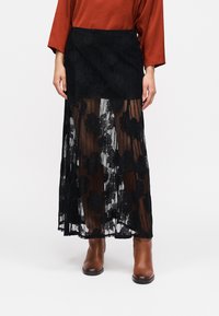 Μακριά φούστα με μαύρη δαντέλα διακοσμημένη με λουλουδάτα σχέδια, μαύρη ζώνη στη μέση, ημιδιαφανές στρώμα και εφαρμοστή σιλουέτα.