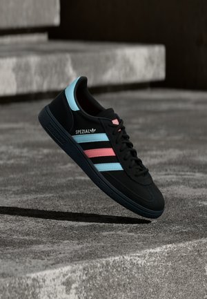 Zwarte Adidas Spezial sneaker met blauwe en roze strepen, zwevend boven een getextureerd grijs betonnen oppervlak met schaduw eronder.