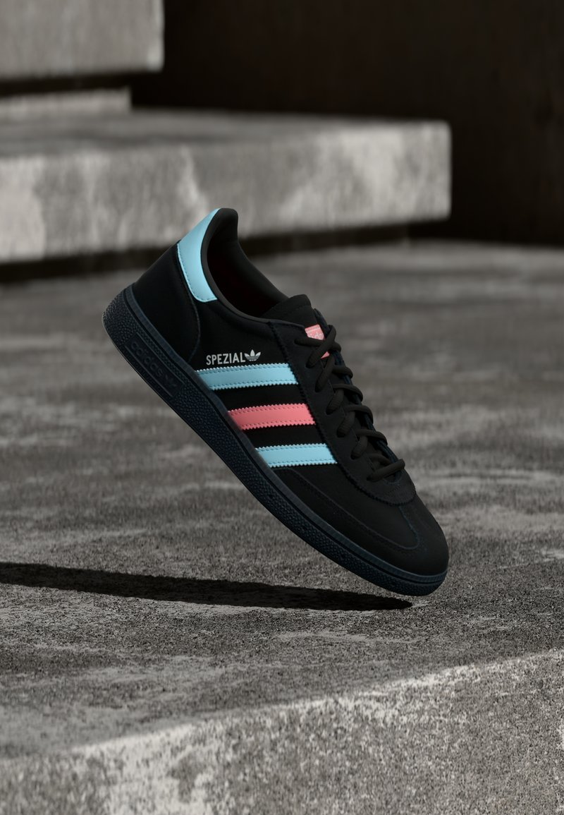 Baskets Adidas Spezial noires avec des rayures bleues et roses, flottant au-dessus d’une surface en béton gris texturé avec une ombre en dessous.
