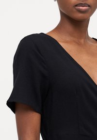 Sort kjole med V-hals, korte ærmer og en tætsiddende overdel. Fremstillet af et glat stof med en let glans. Søm-detaljer synlige.