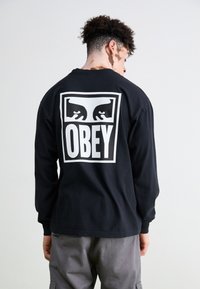 Fekete, hosszú ujjú póló, amelyen hátul egy nagy, fehér "OBEY" logó található, stilizált kezekkel egy téglalap keretben.