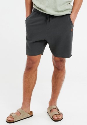 Kurze graue Shorts mit einem elastischen Bund und Kordelzug, aus strukturiertem Stoff und mit Seitentaschen. Getragen mit beigen Sandalen.