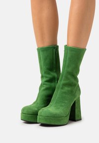 Bottines en daim vertes, à bout arrondi, avec un large talon bloc et des détails de fermeture éclair sur le côté ; texture lisse et coutures minimales.