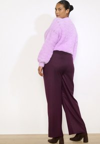 Tumeda juustega naine, kellel on kukk punutud, seljas lavendli värvi kudum, tume lilla laiade jalgadega püksid ja pruunid kontsaga saapad, seisab valge seina ääres.