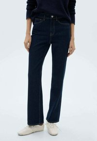 Jeansi din denim albastru închis, cu croială dreaptă, având o talie înaltă, design clasic cu cinci buzunare și cusături galbene contrastante. Asortați cu adidași albi.