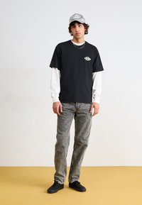 Camiseta de algodón negra sobre una camisa de manga larga blanca, combinada con unos pantalones vaqueros grises desgastados y zapatillas negras. Los complementos incluyen una gorra gris.