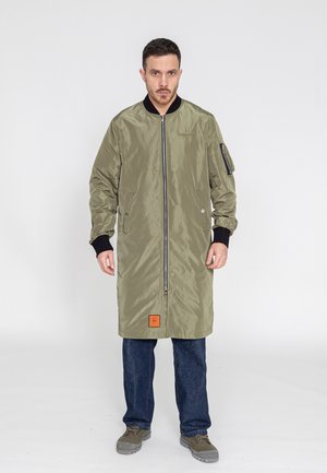 Olivenfarbener langer Bomberjacke mit einem Frontreißverschluss, schwarzen Rippbündchen und einem orangefarbenen Etikett am Saum. Getragen über blauen Jeans und grauen Schuhen.
