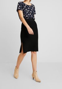 Schwarze, knielange Rock mit einem seitlichen Schlitz, kombiniert mit einer dunkelblauen Bluse mit Blumenmuster. Beigefarbene Stiefeletten mit spitzem Zehenbereich und Absatz.