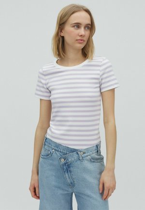 Ung kvinde iført en hvid og lyse lilla stribet t-shirt og asymmetriske lyseblå jeans, står mod en ensfarvet grå baggrund.