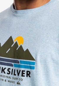 Primer plano de una camisa azul clara con un gráfico de montaña, un sol amarillo y rayas azules sobre el texto "Quiksilver".