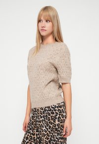 Noor blondide sirgete juustega noor naine kannab beeži kudumisega lühikeste varrukatega sviitrit ja leopardimustrilisi pükse, vaadates külje suunas.