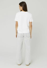 T-shirt en coton blanc à manches courtes, col rond, associé à un jean large gris clair et des chaussures marron, vu de dos.