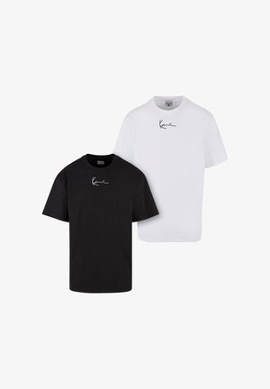 Bărbat purtând un tricou negru cu logo alb tip script pe piept și blugi albastru deschis, stând în fața unui fundal gri simplu.