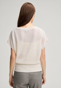 Hellbeige gestrickter Pullover mit kurzen Ärmeln, geripptem Saum und einem leicht transparenten Rückenteil. Weiche Textur, lockere Passform. Getragen mit gemusterten grauen Hosen.