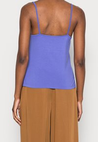 Person som bär en ärmlös lila topp med tunna remmar och lösa bruna byxor, visad från ryggen mot en enfärgad bakgrund.