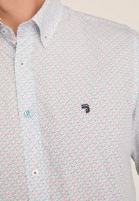 Een button-down shirt in lichte pastelkleuren met een herhalend geometrisch patroon. Voorzien van een klein zwart logo en lichtblauwe knopen.