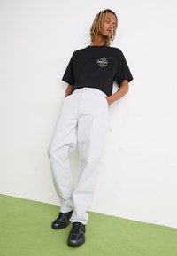 Camiseta negra de gran tamaño con un logo gráfico, combinada con pantalones gris claros de corte relajado y un bolsillo frontal, usados con zapatillas negras.