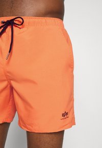Oransje svømmebukser med en glatt tekstur, elastisk livlinning og marineblå snor; har en liten svart logo nederst til venstre.