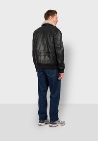 Černá kožená bomber bunda s kožešinovým límcem, pružnými manžetami a pasem, v kombinaci s modrými džíny a sportovními botami. Pohled zezadu.