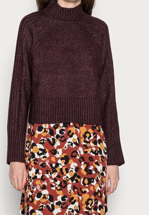 Vrouw die een bordeauxrode geribbelde trui met lange mouwen draagt en een multikleuren luipaardprint rok in bruin, zwart, wit en oranje tinten.