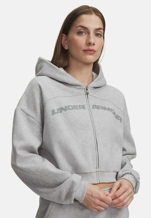 Vrouw die een lichtgrijze zip-up hoodie met het "Under Armour"-logo draagt, staand met de handen bij de tailleband tegen een effen witte achtergrond.