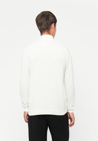 Pull en tricot blanc à manches longues, col haut et poignets côtelés. Texture lisse, coupe décontractée et sans motifs ni accents visibles.