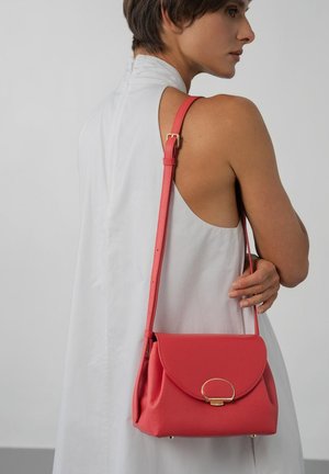 Vrouw in een witte mouwloze jurk met een koraalrode leren handtas met een gouden sluiting en verstelbare riem over haar schouder.