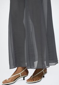 Personne portant un pantalon noir transparent à jambes larges et des sandales blanches à talons bas et brides, montrant les pieds et les jambes inférieures.