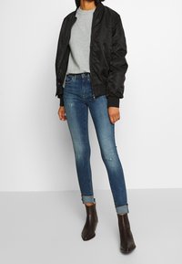 Person klädd i en svart bomberjacka, ljusgrå tröja, blå skinny jeans med uppvikta benen och mörkbruna ankelstövlar som står mot en vit bakgrund.