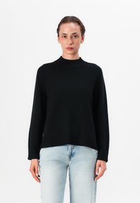 Svart strikket genser med ribbestrikket høy hals, lange ermer og løs passform, samlet med lyseblå jeans for kontrast.