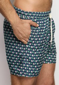 Pantaloni da bagno blu navy con un motivo floreale che presenta fiori rosa e foglie verdi. Vita elastica con cordoncino e dettagli con tasche laterali.