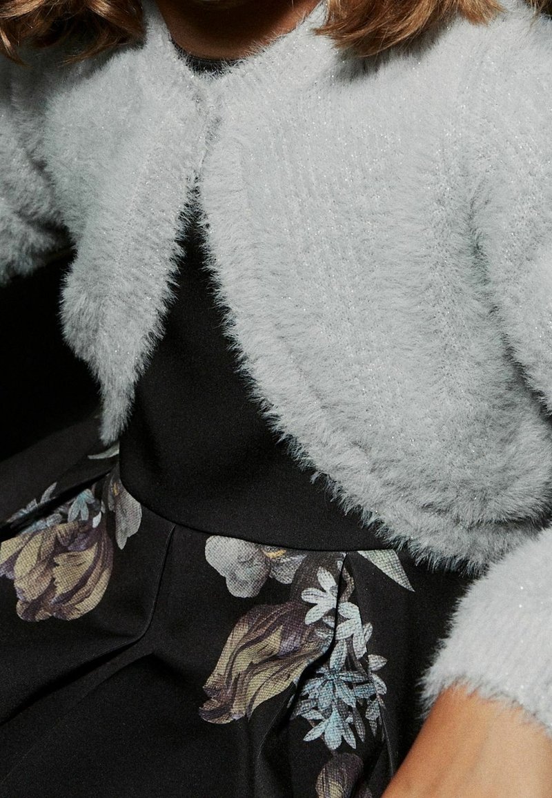 Femme portant un cardigan court et duveteux gris clair par-dessus un haut noir et une jupe noire à motif floral.