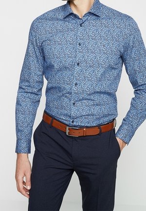 Mann trägt ein eng anliegendes blaues Hemd mit floralem Muster, braunen Ledergürtel und dunkelblaue Hose, eine Hand in der Tasche.