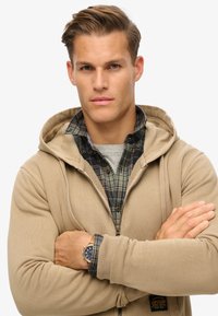 Sudadera con cremallera color beige sobre una camiseta gris y una camisa de franela de cuadros, con capucha ajustable, puños de canalé y un parche de logo. Reloj marrón en la muñeca.