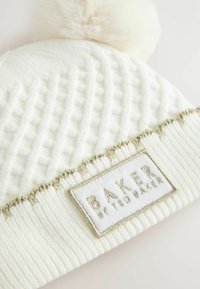 Weißer gestrickter Mütze mit strukturiertem Muster, ausgestattet mit einem flauschigen Pompon und einem dekorativen Band mit goldenen Akzenten, das ein Etikett mit der Aufschrift "BAKER" trägt.