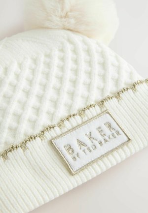 Weißer gestrickter Mütze mit strukturiertem Muster, ausgestattet mit einem flauschigen Pompon und einem dekorativen Band mit goldenen Akzenten, das ein Etikett mit der Aufschrift "BAKER" trägt.