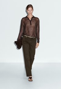 Chemise longue en voile marron à manches longues avec un col, pantalon droit vert olive, et sandales noires à lanières. Accessoire à la main : un sac à main foncé.