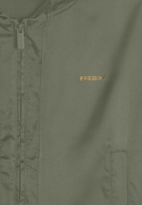 Chaqueta verde con cremallera y textura suave, que presenta un pequeño logo dorado "FREDDY" en el lado izquierdo del pecho y bolsillos laterales.