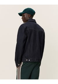 Giacca in denim scuro con colletto classico, dettagli di cucitura a vista e bottoni metallici, abbinata a un cappello in velluto a coste verde e pantaloni a righe.