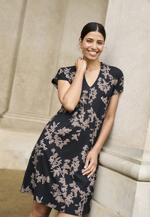 Femeie cu păr închis la culoare, purtând o rochie neagră cu modele florale bej, zâmbind și sprijinindu-se de o coloană de piatră bej.