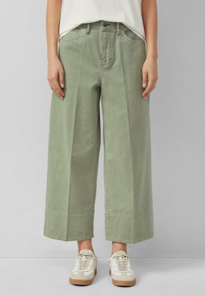 Osoba nosząca szerokie, skrócone jasnozielone spodnie z przednimi kieszeniami i białe trampki na jednolitym tle.